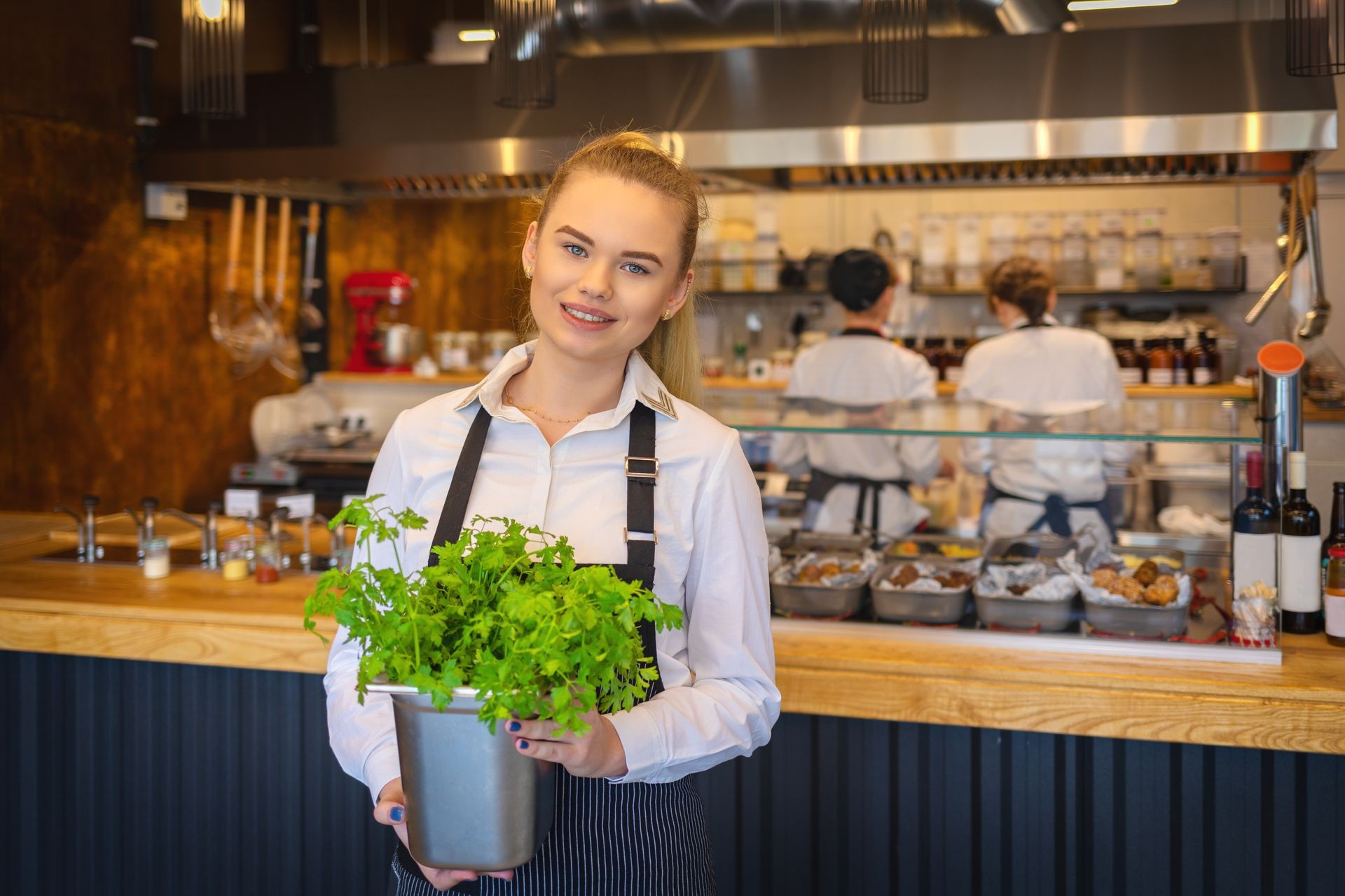 Happy restaurant owner at counter holding pot with fresh parsley with two waitress working in background – Smiling chef restaurant woman cooking with fresh ingredients smiling to camera